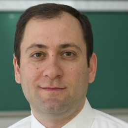 dark-haired male professor in white, collared shirt and tie, standing in front of a green chalkboard